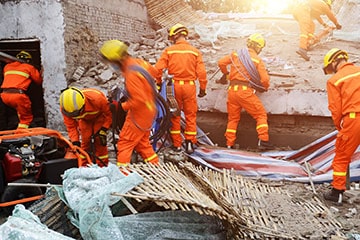 six emergency workers in hard hats and protective jumpsuits search through ruined building