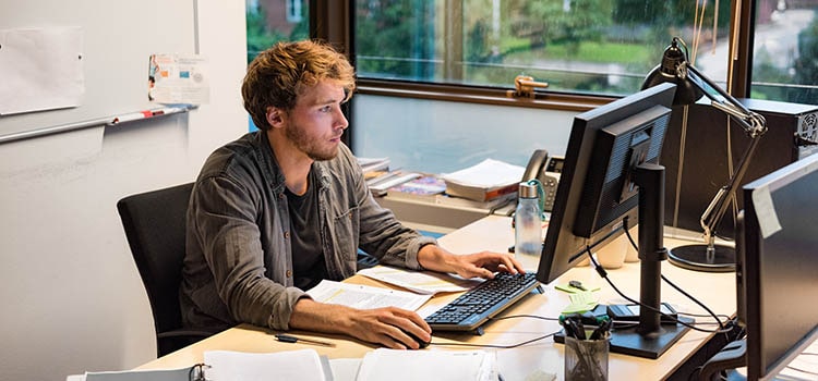 phd candidate works at desk from spread out notes and whiteboard in background