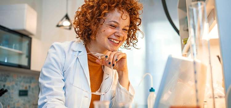 smiling person sits at work bench looking at laptop screen while wearing lab coat and holding safety glasses