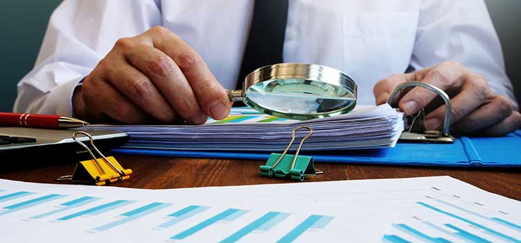 hands hold magnifying glass pore over stack of financial documents