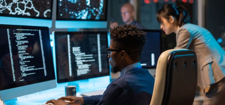 cyber security specialist sitting at a desk observes multiple computer monitors with a colleague