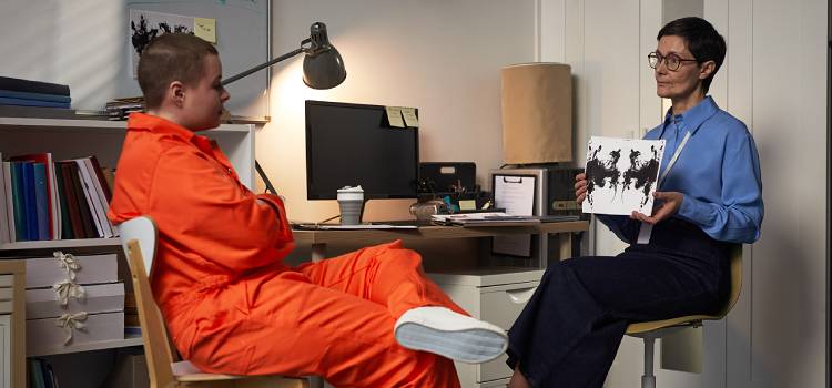 Caucasian teenager sitting in orange prison jumpsuit facing middle aged Caucasian woman therapist holding inkblot test card during psychological evaluation in office setting